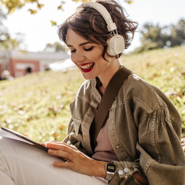 Lovely woman in jacket and headphones sitting on grass outdoors. Happy girl with curly hairstyle holding smartphone outside..